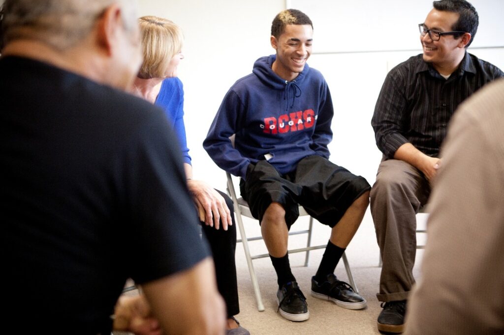 Smiling young man surrounded by adults sitting in chairs giving encouraging smiles. 